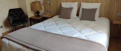 Una cama bien hecha con colcha y almohadas color beige, un cabecero de madera, una mesita de noche con una lámpara y un papel tapiz floral.