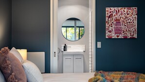 Un dormitorio con una cama, una lámpara de noche, un espejo redondo, una ventana y una pared con un cuadro colorido.