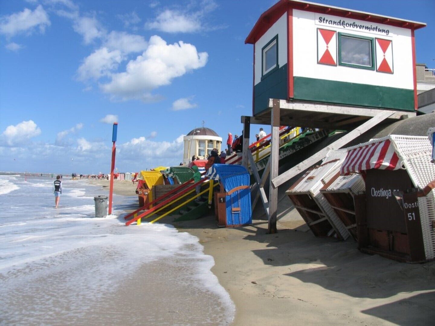 Una torre de salvavidas con diseño rojo y blanco, una escalera que conduce a ella, y una playa al fondo.