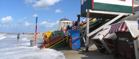 Una torre de salvavidas con diseño rojo y blanco, una escalera que conduce a ella, y una playa al fondo.