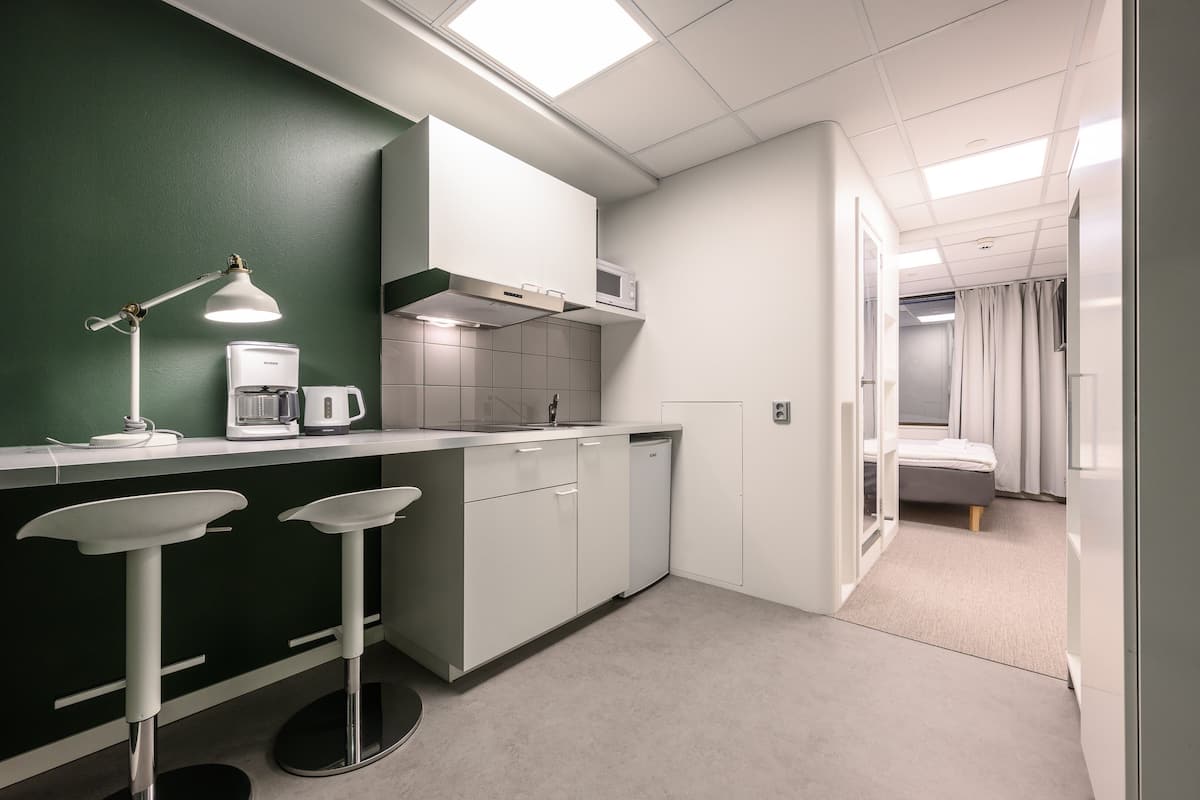 Een kleine keuken met een groene accentmuur, witte kasten, een magnetron en een koffiezetapparaat. Er staan twee barkrukken bij een klein keukentje.
