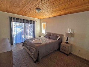 A bedroom with a wooden ceiling, a bed with grey bedding, a nightstand with a lamp, and a window with curtains. - Waterfront Home on Double Lot - Boat Dock, Hot Tub and More! (South Lake Tahoe)