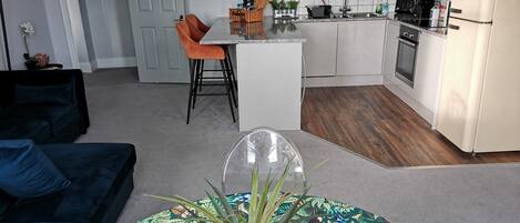 A modern kitchen with white cabinets, a built-in oven, and a white refrigerator. There is a dining area with a white table and orange chairs. A potted plant is on a table in the foreground.