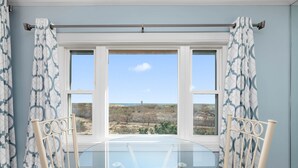 Una habitación con mesa y sillas de vidrio, una ventana con vista a la playa y cortinas estampadas.
