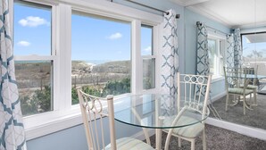 Un comedor con mesa y sillas de vidrio, ventanas amplias con cortinas, y vista a un paisaje.