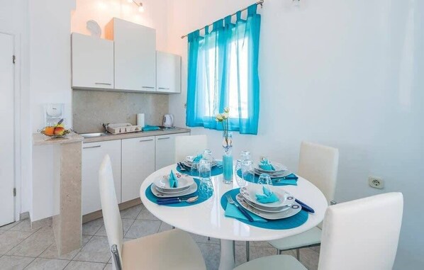 Une cuisine moderne avec des meubles blancs, une table à manger dressée pour quatre personnes avec des serviettes et de la verrerie bleues, et une fenêtre ornée de rideaux bleus.