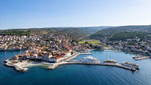 Vue aérienne d’une ville côtière avec un port de plaisance, des maisons et un paysage vallonné en arrière-plan.