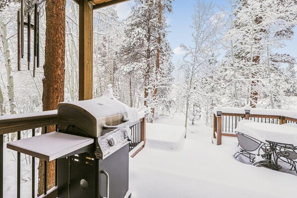 A wooden deck with a grill and a table covered in snow, surrounded by snow-covered trees.