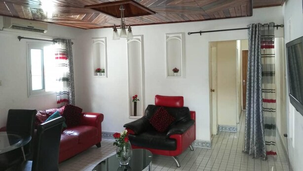 Una sala de estar con un sofá rojo, un sofá negro y una mesa de centro de vidrio. Hay dos ventanas con cortinas, una puerta y una araña.