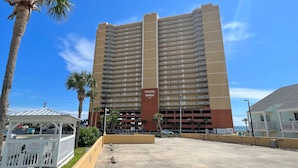 Un hotel de gran altura con varios balcones, un letrero vertical central y un estacionamiento con palmeras.