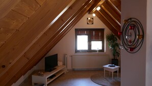 Ein gemütliches Dachzimmer mit geneigter Decke, einem Heizkörper, einem Fenster mit Jalousien, einem kleinen Tisch, einem Fernseher auf einem Regal und einem Blumentopf.