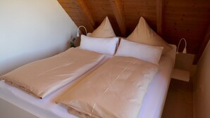 Ein Bett mit beigefarbener Bettwäsche, ein weißer Nachttisch und eine Holzbalkendecke.