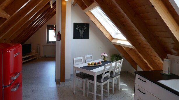 Ein Essbereich mit einem weißen Tisch, Stühlen und einer Obstschale. Links ist ein rotes Gerät zu sehen und ein Fenster mit Blick ins Freie.