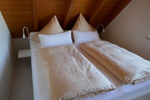 A neatly made bed with white linens and two pillows, a wooden ceiling, and a white bedside table with a lamp.