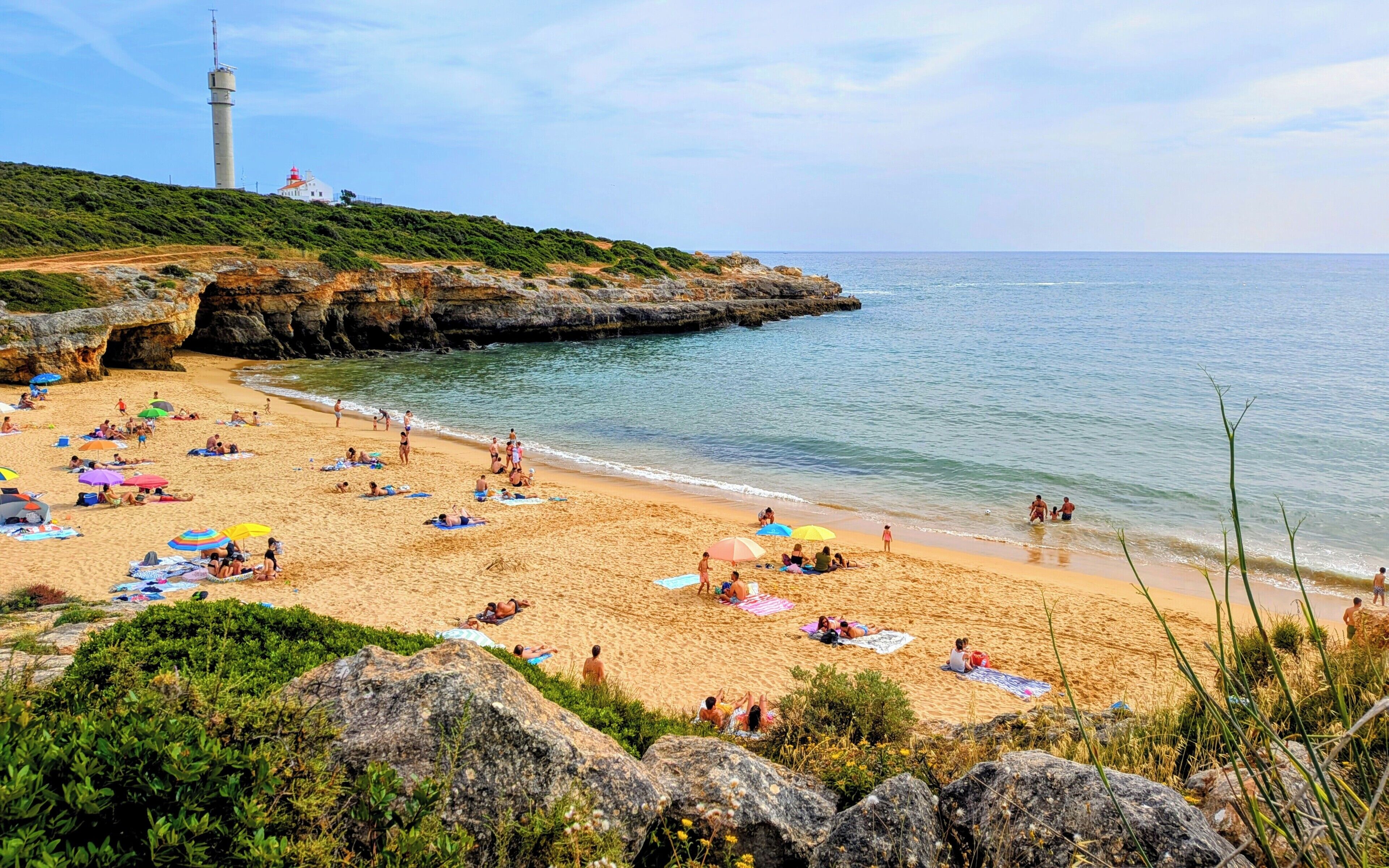 A beach with people sunbathing, umbrellas, and a lighthouse in the background.