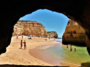 A beach scene viewed through a cave opening, with people on the sand and in the water, and cliffs in the background.