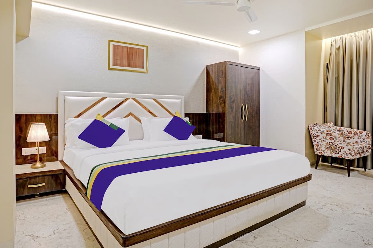 A neatly made bed with a headboard, a nightstand with a lamp, a wardrobe, and a chair with a floral pattern.