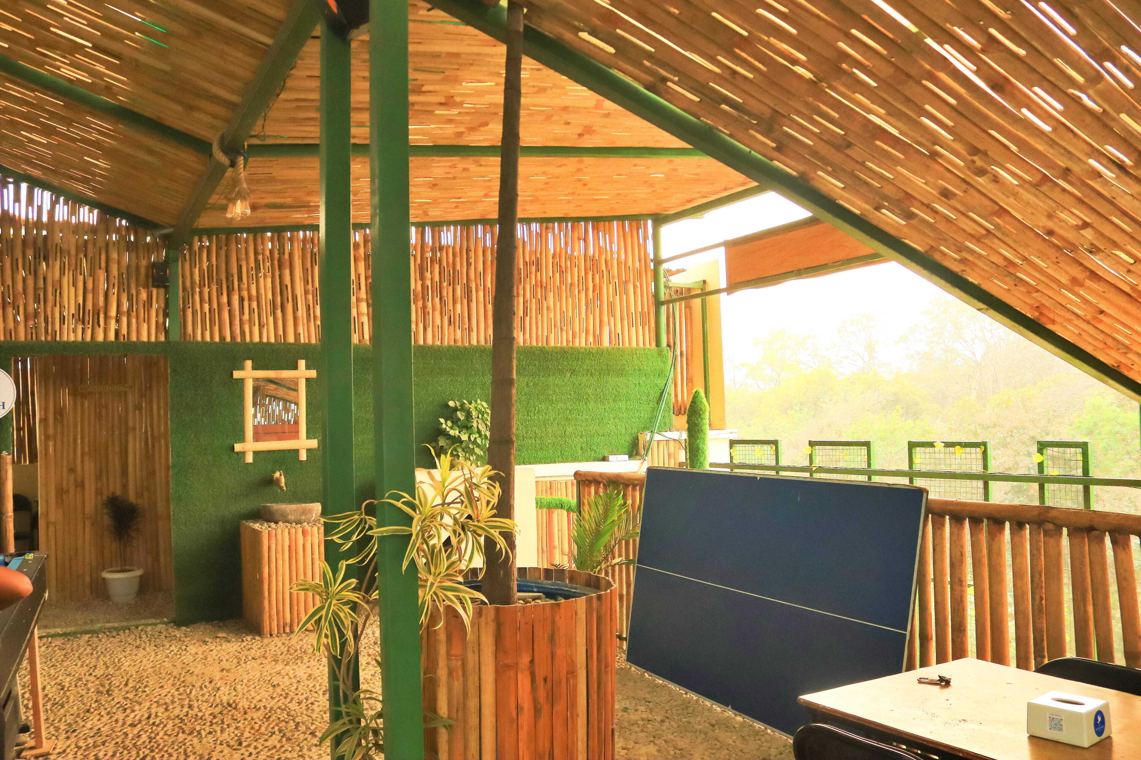 A room with a sloped ceiling, bamboo walls, a green artificial grass floor, a wooden table with a chair, and a window with a view of trees.