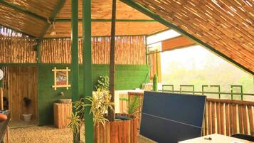 A room with a sloped ceiling, bamboo walls, a green artificial grass floor, a wooden table with a chair, and a window with a view of trees.