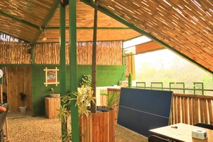 Una habitación con techo inclinado, paredes de bambú, suelo de césped artificial verde, una mesa de madera con una silla y una ventana con vistas a árboles.