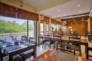 A restaurant with multiple tables and chairs, large windows offering a view of the street, and a ceiling fan. - Hotel Peaceland Lumbini (Lumbini)