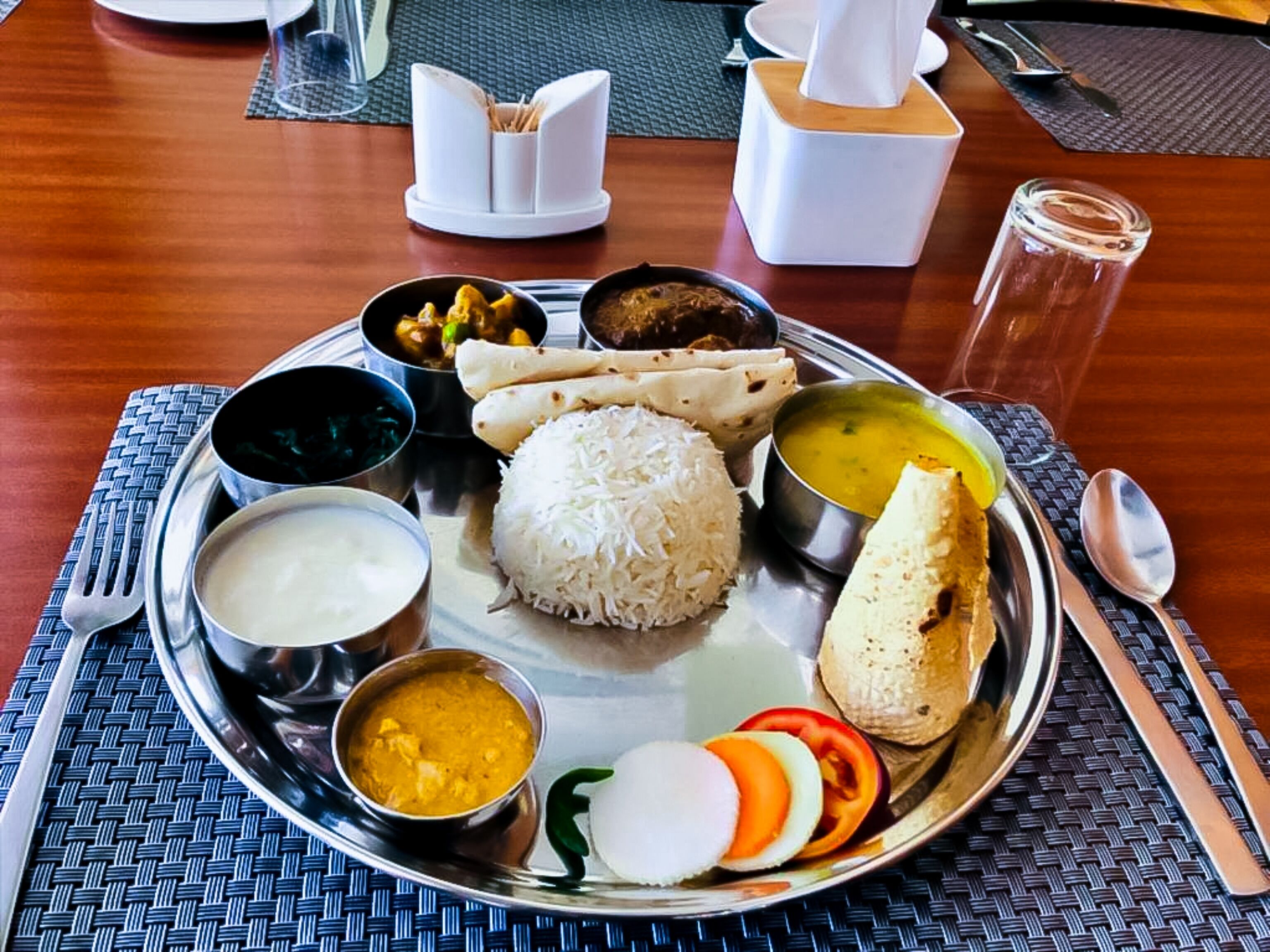 A traditional Indian meal on a metal plate with rice, various curries, flatbreads, and a slice of tomato.