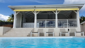 Uma casa moderna com piscina, apresentando um alpendre coberto com cadeiras e uma escadaria que leva à entrada.