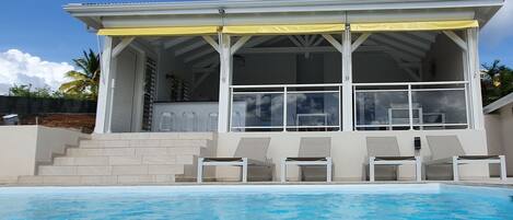 A modern house with a pool, featuring a covered patio with chairs and a staircase leading to the entrance.