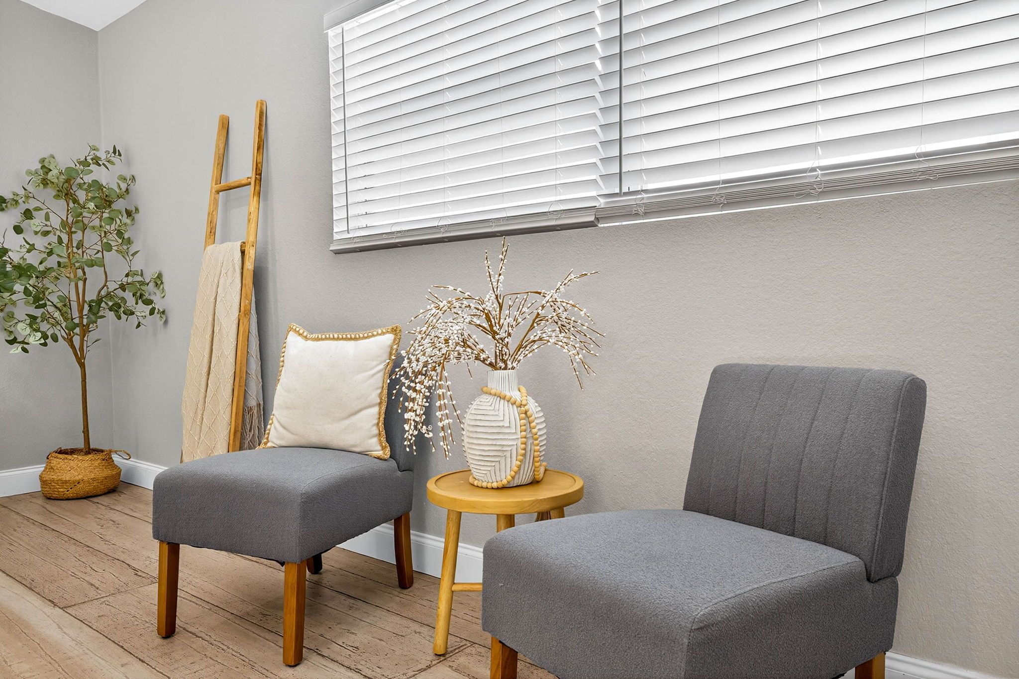Une pièce comprenant deux fauteuils gris à revêtement en tissu, une petite table ronde en bois, un panier tressé, une grande échelle en bois et une plante en pot.