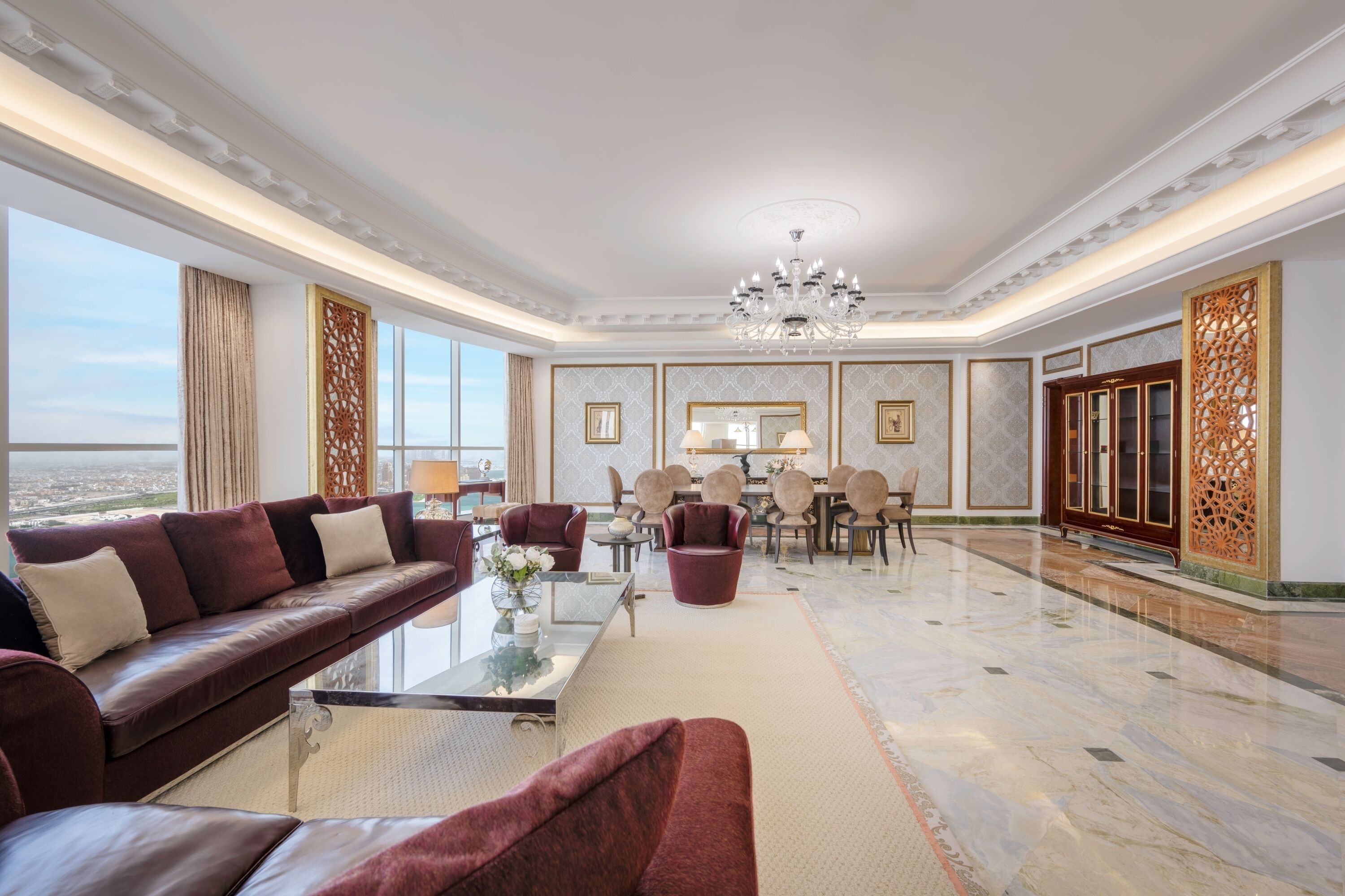 A spacious living room with a large sofa, a coffee table, and a chandelier. There is a dining area with chairs and a table in the background.