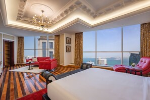 A hotel room with a large bed, red armchairs, a red sofa, a small table, and a view of the city and sea. - Dusit Hotel & Suites Doha (Doha)