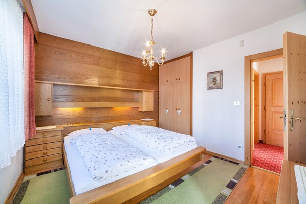 Quarto com cabeceira de madeira, um candelabro, cama com roupa de cama floral, guarda-roupa de madeira e porta para outro cômodo.