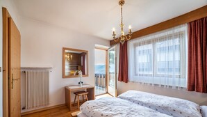 Ein Schlafzimmer mit einem Holztisch, einem Bett mit floralem Bettbezug, einem Kronleuchter und Blick durch ein Fenster mit Vorhängen.
