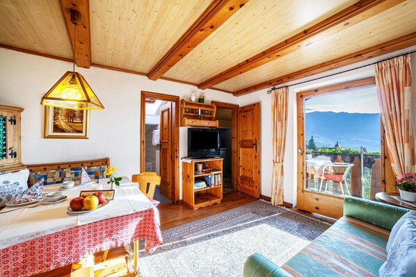 A cozy room with a wooden ceiling, a television, a dining table with a red tablecloth, and a view of the mountains through a glass door.