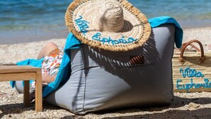 Escena de playa con un sombrero de paja y un cojín bordado con la inscripción 'Villa Euphorio'.