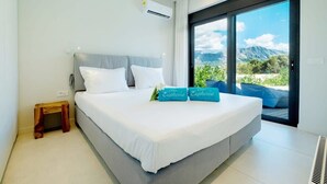 Un dormitorio moderno con una cama grande, dos almohadas, una mesita de noche y un taburete de madera pequeño. Se observa una ventana amplia con vista a la montaña.