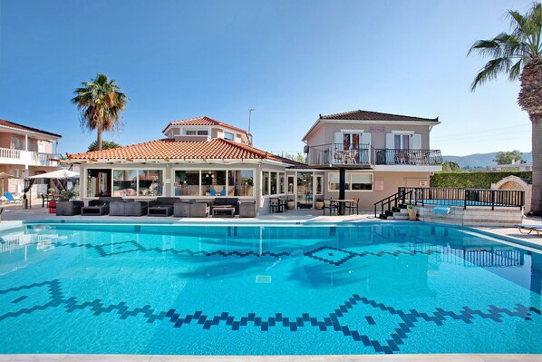 Une piscine dont le fond est décoré de motifs géométriques en mosaïque, un espace extérieur aménagé avec un toit, et un bâtiment doté d’un balcon.