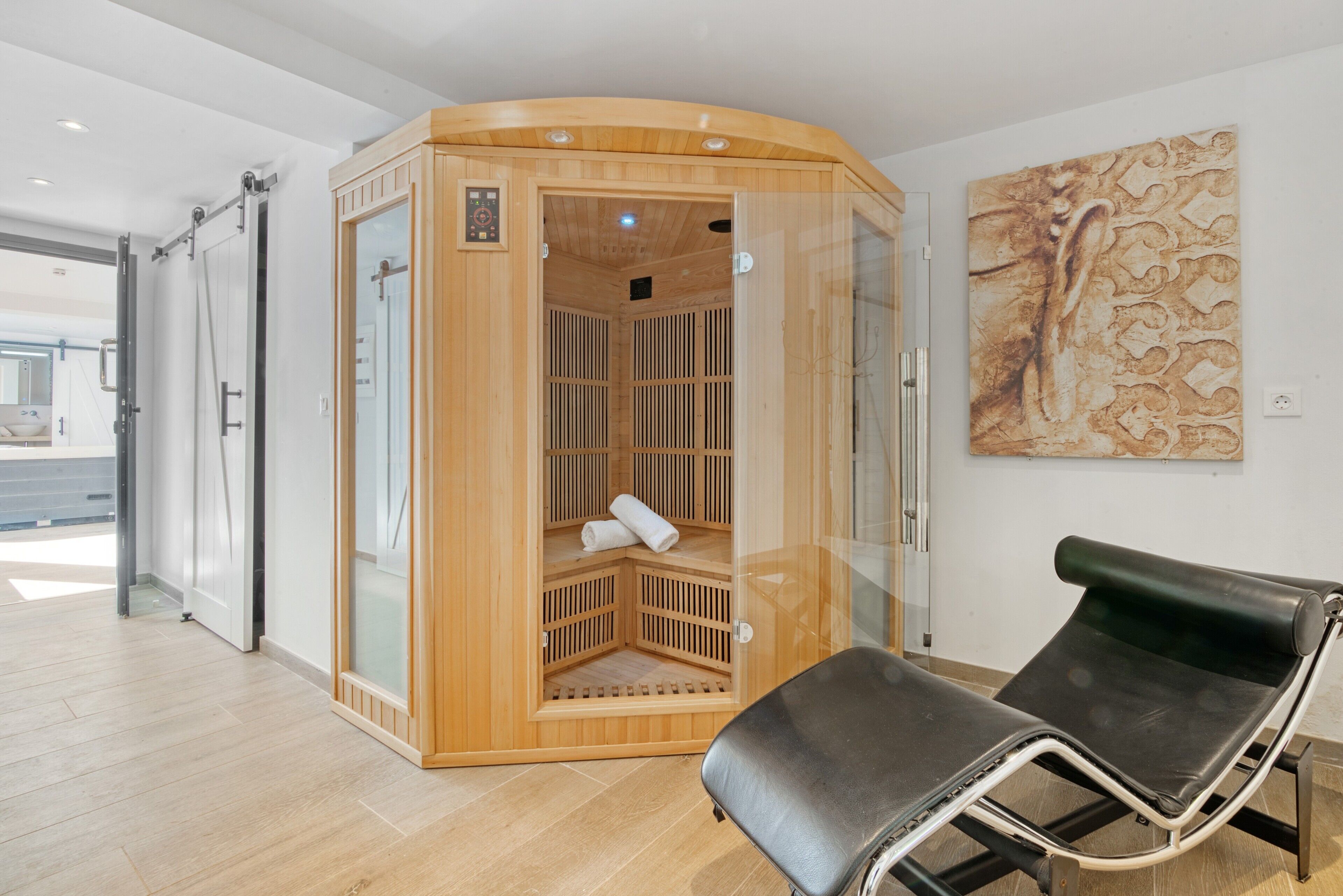 Un sauna avec une structure extérieure en bois, une porte en verre et un fauteuil noir placé au premier plan.