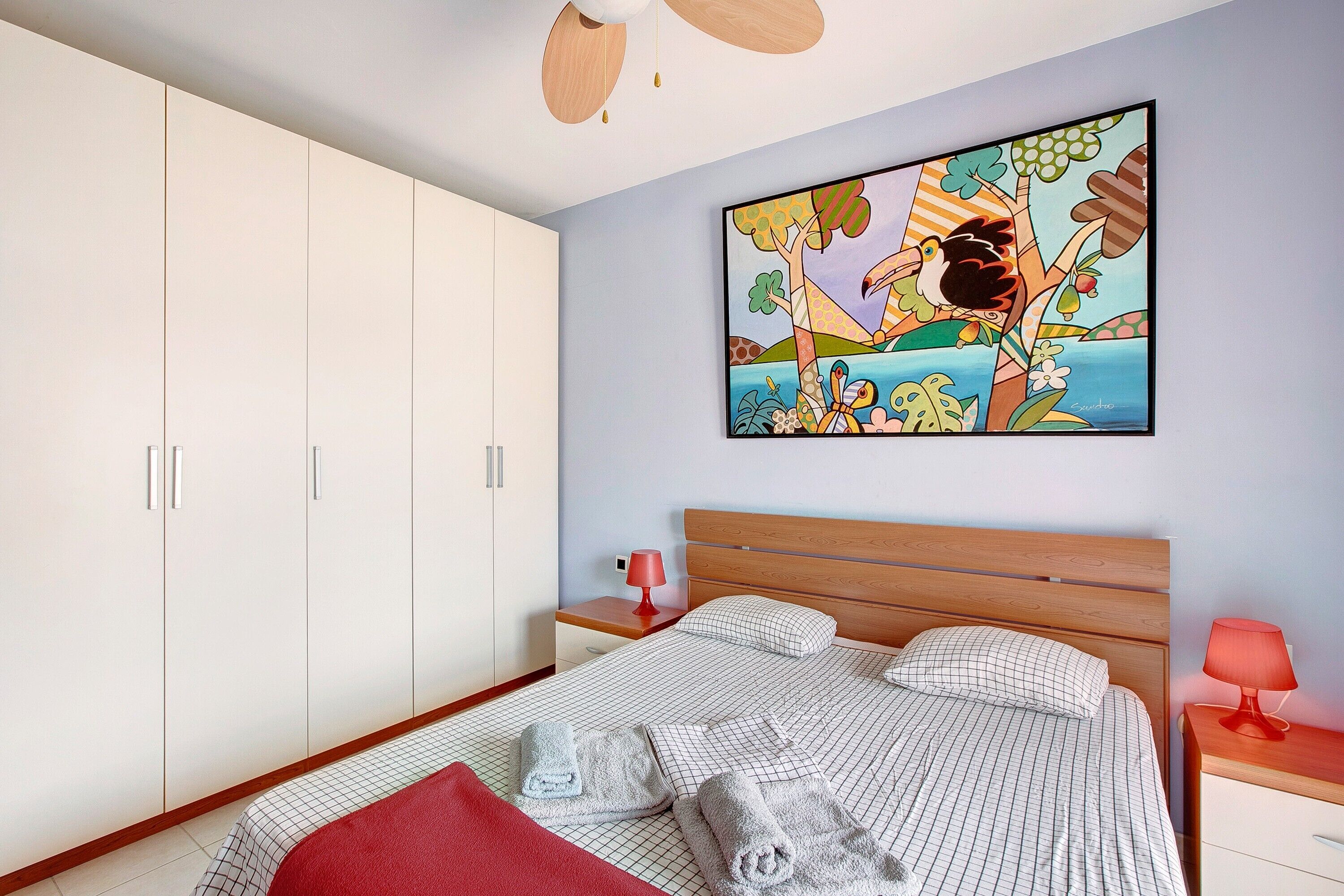 Een slaapkamer met een bed, een garderobe, een nachtkastje, een lamp en een schilderij aan de muur.