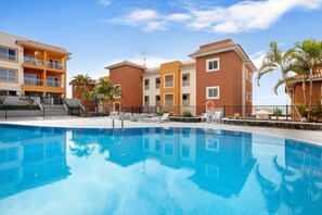 A swimming pool with a clear blue surface, surrounded by a building with balconies and a tiled roof.