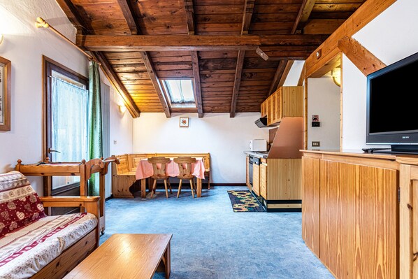 Una habitación acogedora con techo de madera, un sofá, una mesa de comedor con sillas, una zona de cocina con microondas y un televisor.