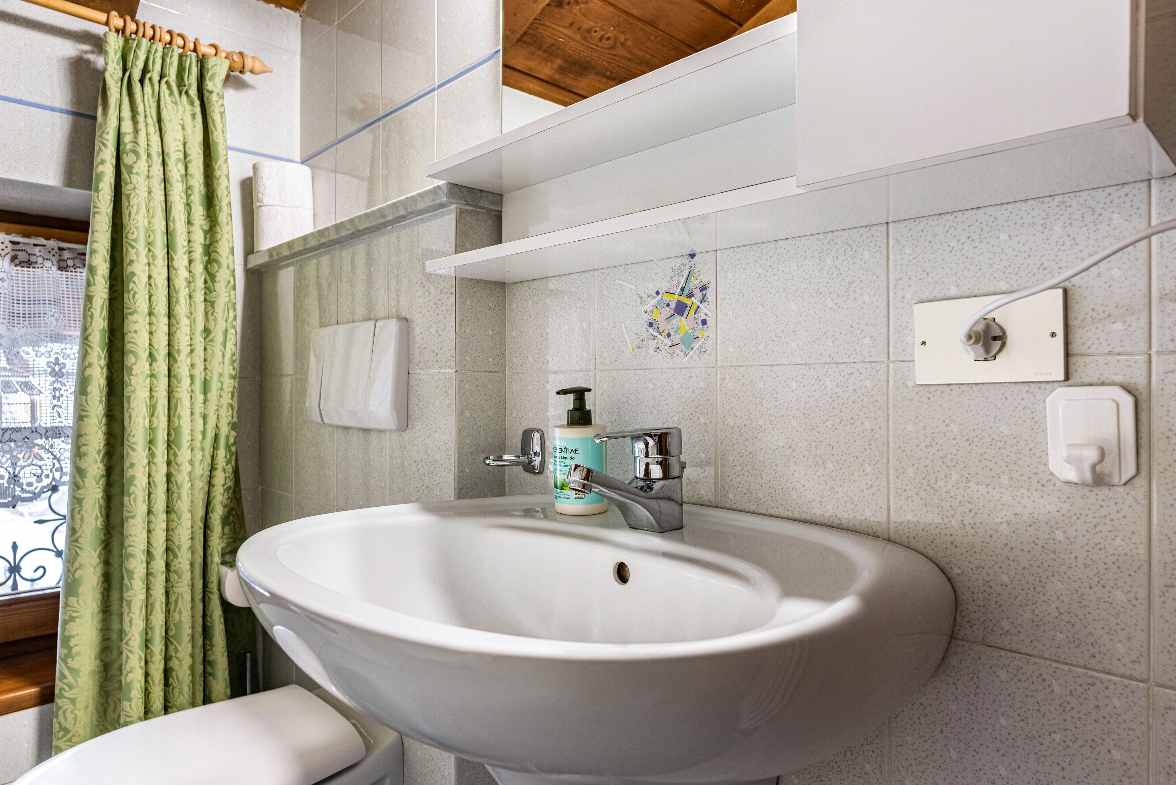 Une salle de bain avec un lavabo blanc, un rideau de douche à motifs verts et un flacon de savon liquide fixé au mur.