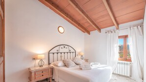 Ein Schlafzimmer mit einem Bett, Nachttischlampen, einem Schminktisch, einem Fenster mit Vorhängen und einer Holzbalkendecke.