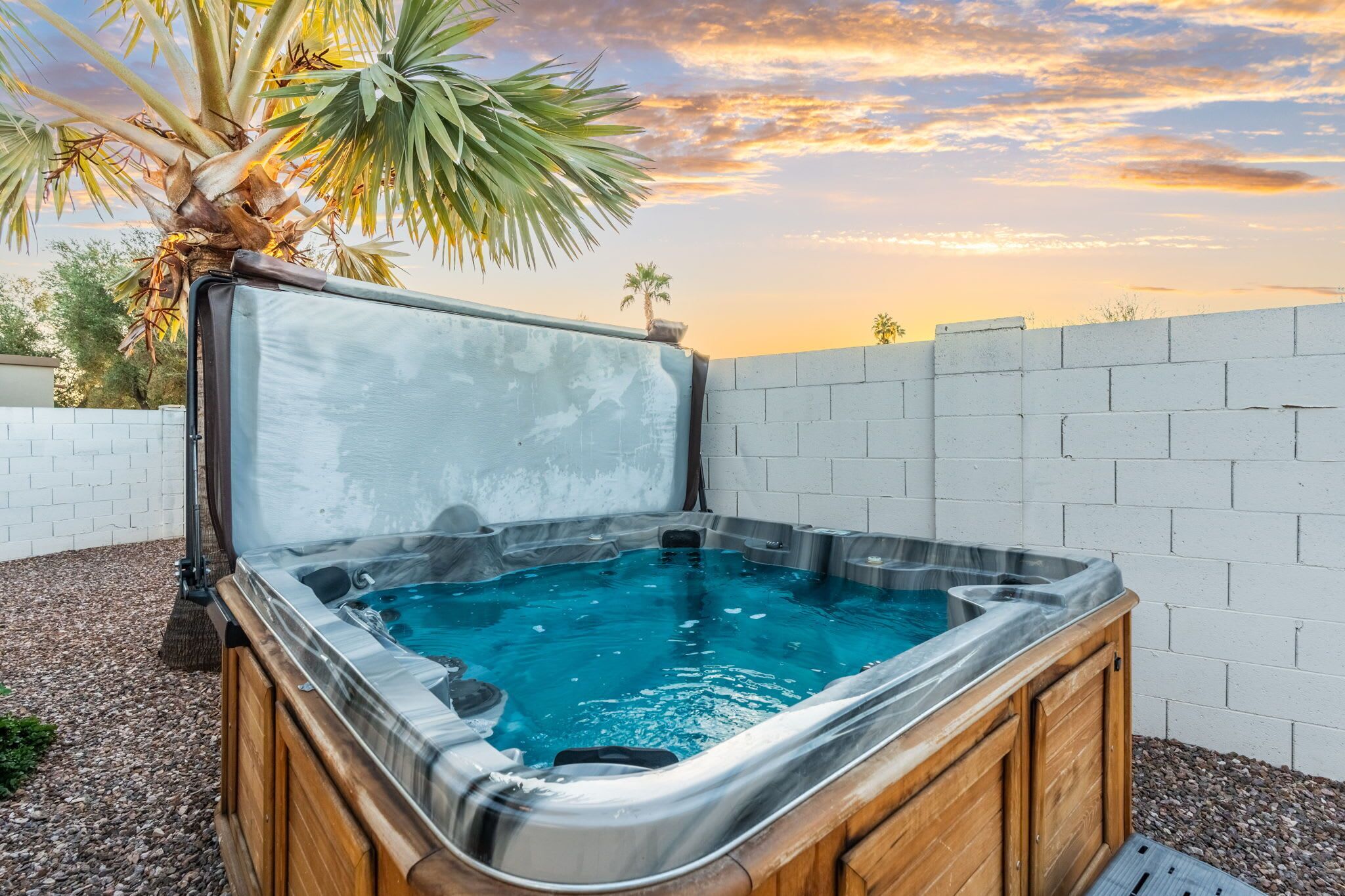 A hot tub with a wooden frame and a clear cover, surrounded by a white brick wall and a palm tree.