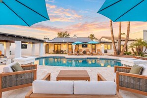 An outdoor sofa and armchairs sit under blue umbrellas facing the pool, with fire bowls on each side and the house lit up in the back. - Desert Playground |Pool|Sportcourt |Bocce | Cabana (Scottsdale)