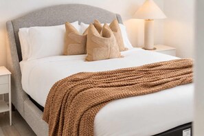 Ein ordentlich bezogenes Bett mit grauem Kopfteil, weißer Bettwäsche und einer braunen Strickdecke.