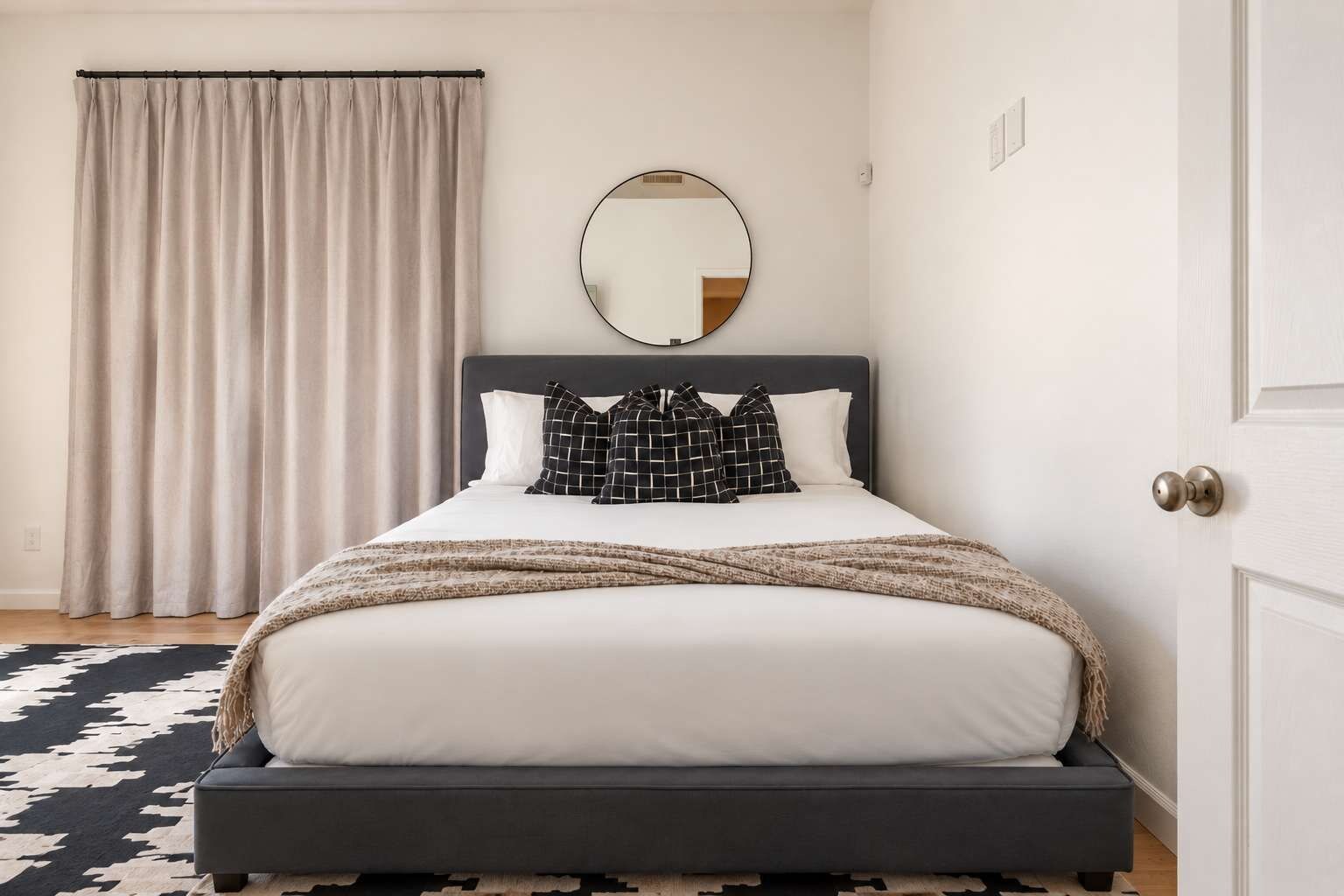 Un dormitorio con una cama, un espejo redondo, una puerta y una alfombra estampada.