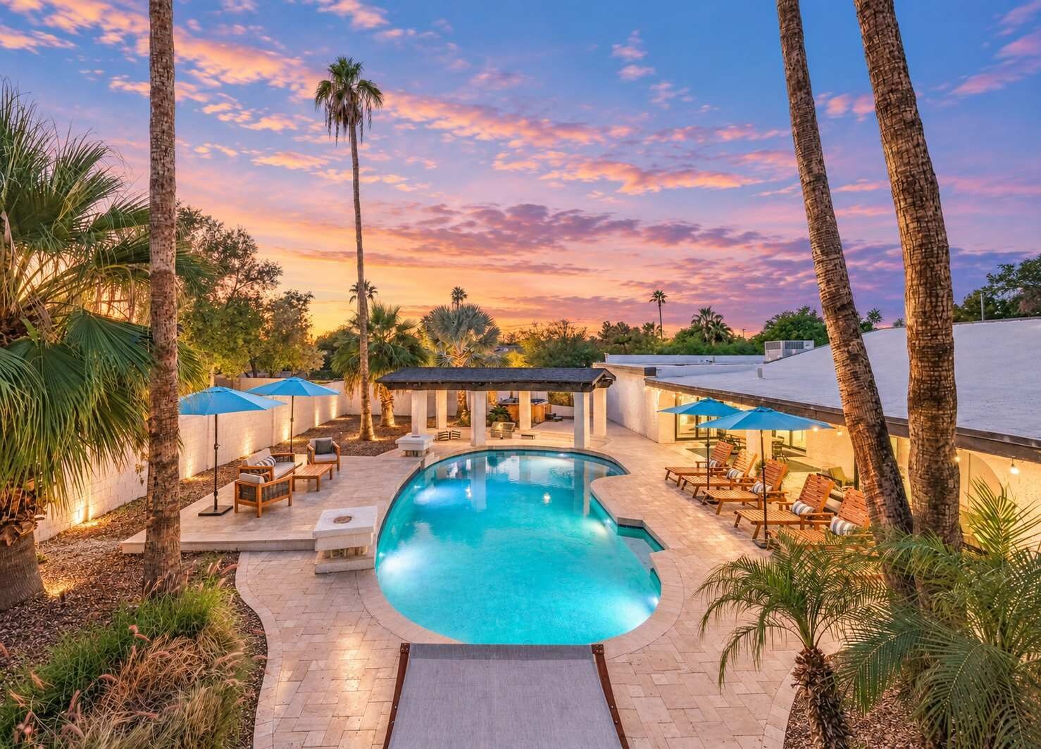 A large lit pool sits at the center of the backyard, with lounge chairs, blue umbrellas, a covered pavilion, and tall palms all around.