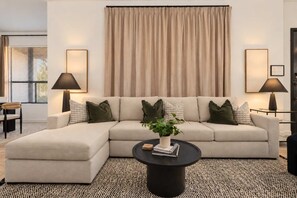 Un salon moderne comprenant un canapé d’angle, deux tables d’appoint avec des lampes, une table basse avec une plante et une grande fenêtre avec des stores.