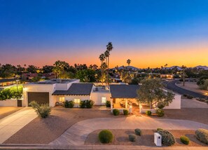 A single-story home with a circle driveway, a lit front porch, and palm trees all around, with mountain views on the skyline at sunset. - Desert Playground |Pool|Sportcourt |Bocce | Cabana (Scottsdale)